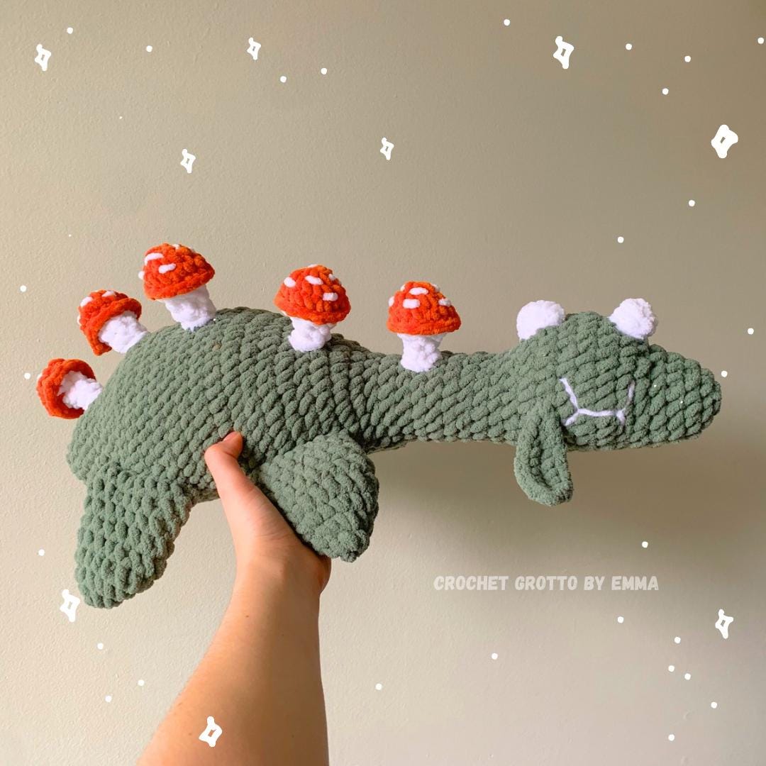 A sage green crochet Nessie plushie with red amanita mushrooms sprouting on its back.