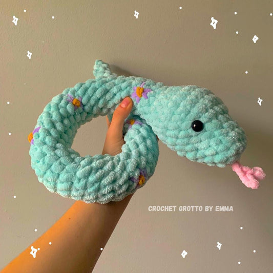 A turquoise, fluffy crochet snake plushie with embroidered flower details (in purple).